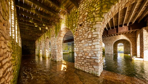 A stone archway structure with water flowing beneath, featuring moss-covered walls and wooden beams above.