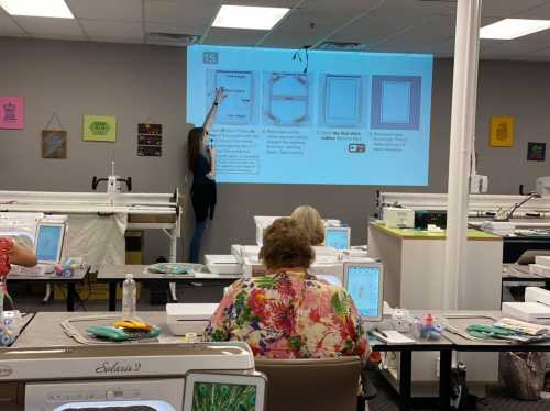 A woman points at a presentation screen while attendees work on sewing machines in a classroom setting.