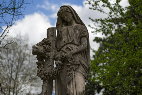 A weathered statue of a woman holding flowers, standing beside a cross, with trees and a cloudy sky in the background.