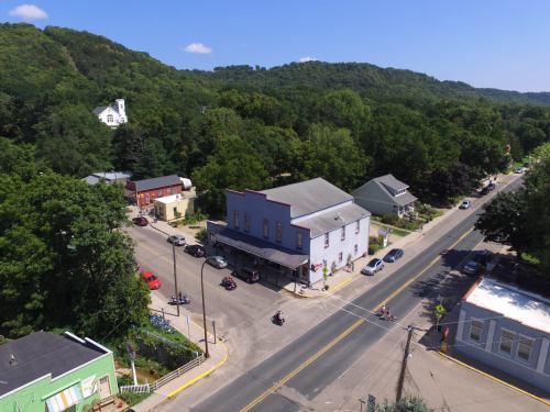 Aerial view of a small town with green hills, a church, and a few buildings along a street, with motorcycles parked nearby.