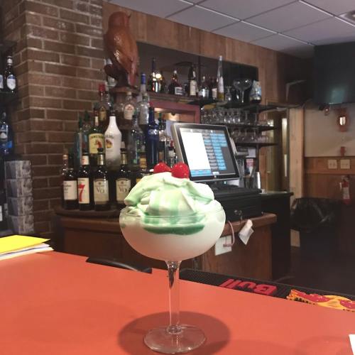 A colorful dessert in a glass, topped with whipped cream and cherries, sits on a bar counter with bottles in the background.