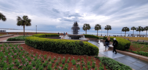 A scenic park with a fountain, surrounded by greenery and flowers, under a cloudy sky, with people walking nearby.