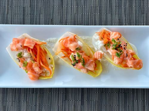 Three sushi-style bites on a white plate, featuring salmon, rice, and carrots on endive leaves, garnished with green onions.