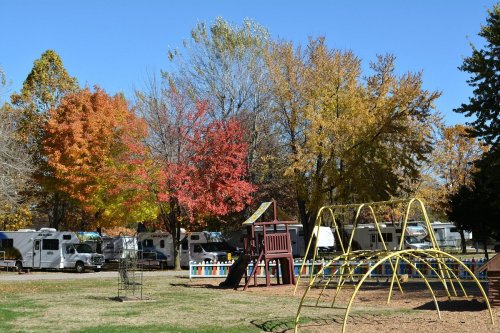 A playground with climbing equipment surrounded by colorful autumn trees and parked RVs in the background.