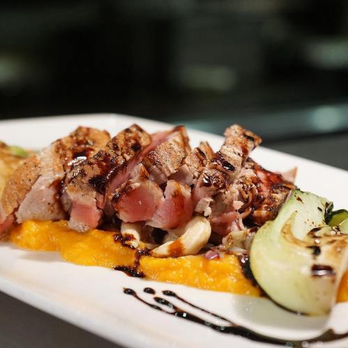 Grilled sliced meat on a bed of mashed squash, garnished with vegetables and a drizzle of sauce.
