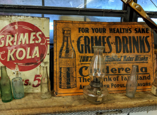 Vintage signs for Grimes Kola and Grimes Drinks, with an old lantern and glass bottles on a wooden shelf.