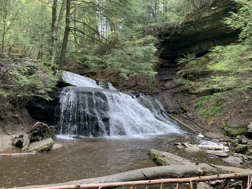 A serene waterfall cascades over rocks into a calm pool, surrounded by lush green trees and a rocky landscape.