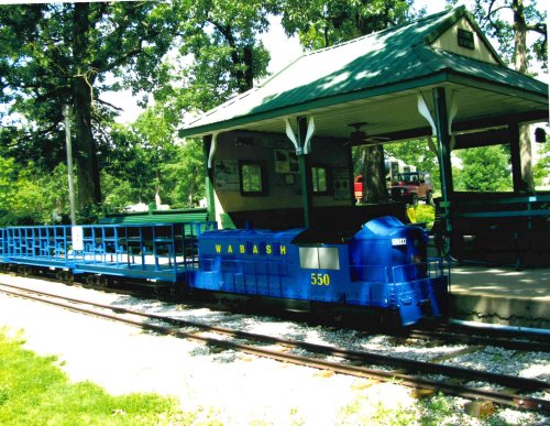 A blue miniature train labeled "Wabash 550" parked at a green-roofed station surrounded by trees.