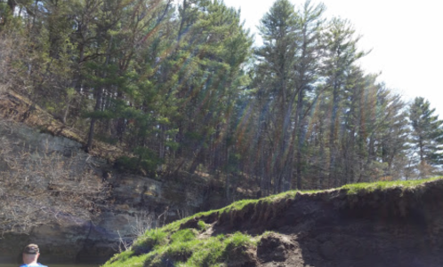 A person stands near a rocky shore with trees and a bright sky in the background, capturing a serene natural landscape.