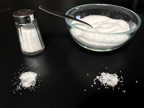 A glass bowl of white sugar with a spoon, next to a salt shaker and two small piles of sugar on a dark surface.