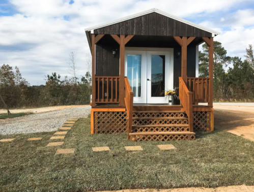 A small, dark wooden cabin with a porch, surrounded by gravel and grass, under a cloudy sky.