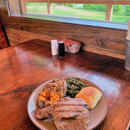 A plate with grilled pork chop, stuffed baked potato, green beans, and a roll, set on a wooden table with a view outside.