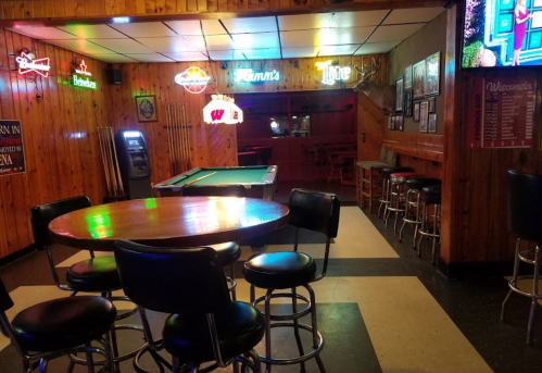 A cozy bar interior with wooden walls, neon signs, a pool table, and a round table with black stools.