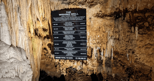 A sign in a cave listing donations for a wishing well, surrounded by stalactites and rock formations.