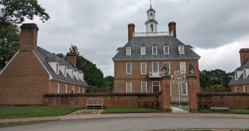 Historic brick mansion with a central tower, surrounded by manicured lawns and a decorative gate, under a cloudy sky.