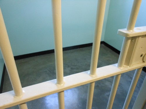 View of a jail cell with white bars and a concrete floor, featuring light blue walls.