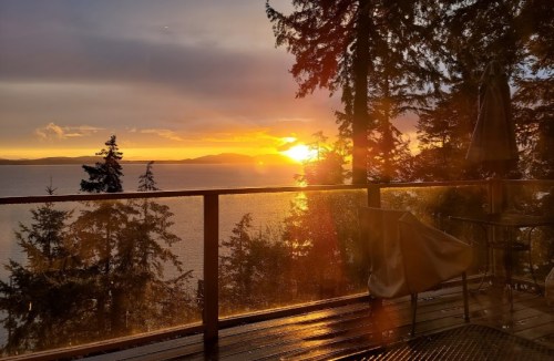 Sunset over a tranquil lake, framed by trees and a wooden deck with a table and chairs.