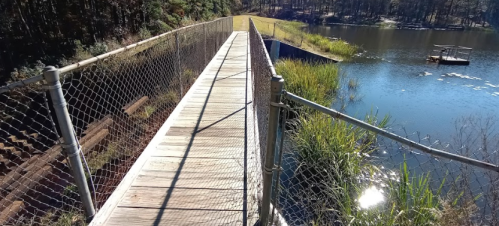 A wooden bridge with a chain-link fence spans a calm pond, surrounded by greenery and trees. Sunlight reflects on the water.