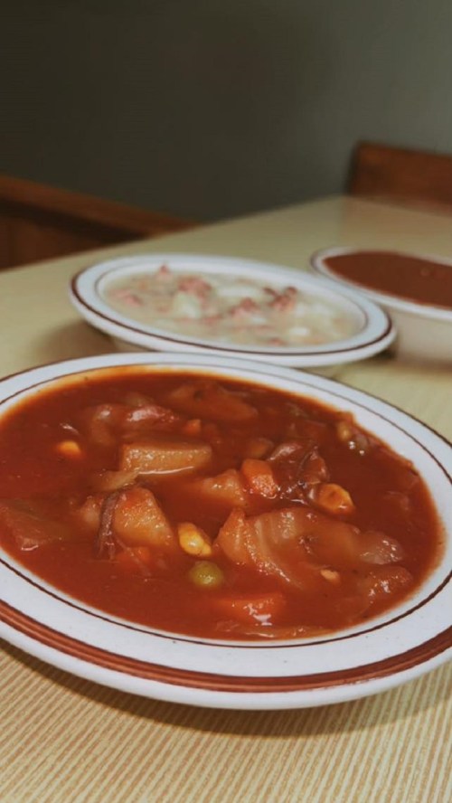 A close-up of a bowl of thick red soup with vegetables, with two other bowls of soup in the background.