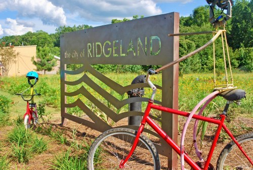 A sign for the city of Ridgeland with decorative bicycles and a colorful helmet in a grassy area.