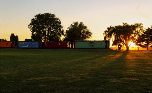 A row of colorful train cars silhouetted against a sunset, with trees and grass in the foreground.