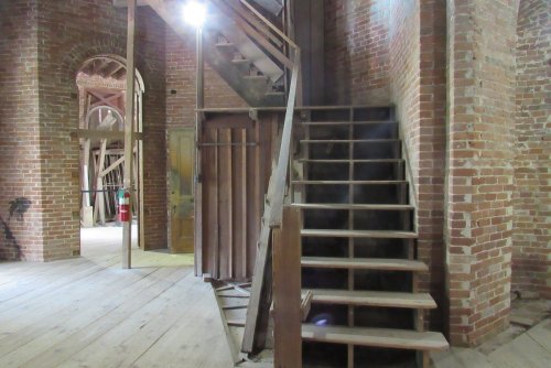 Interior of a brick building featuring a wooden staircase and a doorway, with exposed beams and wooden flooring.