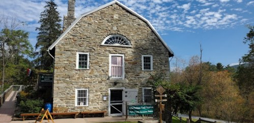 A stone building with a large window and wooden benches outside, surrounded by trees and a blue sky.