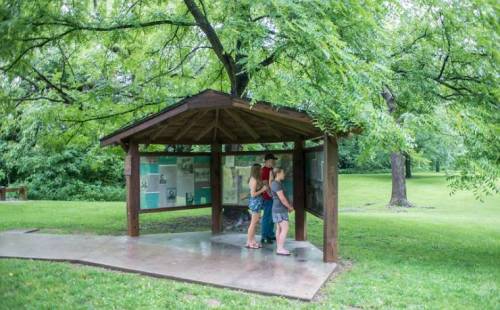 Two people stand under a wooden shelter, reading informational displays in a green park surrounded by trees.
