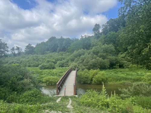 A wooden bridge spans a calm river, surrounded by lush greenery and a cloudy blue sky.