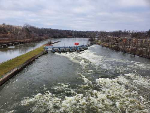 A river with a dam and flowing water, surrounded by trees and buildings under a cloudy sky.