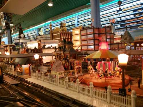 A detailed miniature train setup featuring a carousel, buildings, and various decorative elements under bright lights.