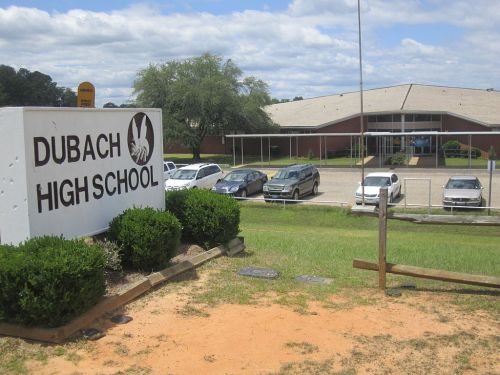 Sign for Dubach High School with a view of the school building and parked cars in a grassy area under a cloudy sky.