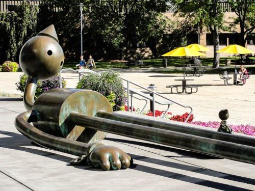 A large bronze sculpture of a whimsical cat lying on the ground in a park with picnic tables and yellow umbrellas nearby.
