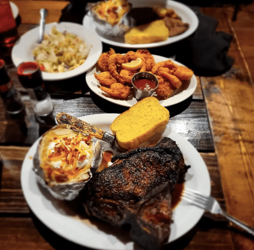 A hearty meal featuring a grilled steak, baked potato, shrimp, coleslaw, and cornbread on a wooden table.