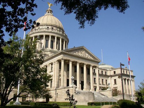 A grand government building with a large dome, columns, and flags, surrounded by trees and a clear blue sky.