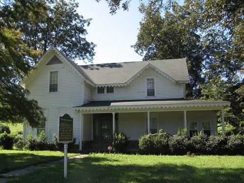 A white, two-story house with a porch, surrounded by greenery and a historical marker in the foreground.