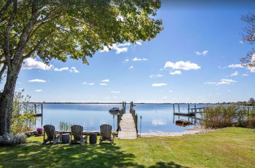 A serene lakeside view with a wooden dock, chairs, and a clear blue sky dotted with clouds.