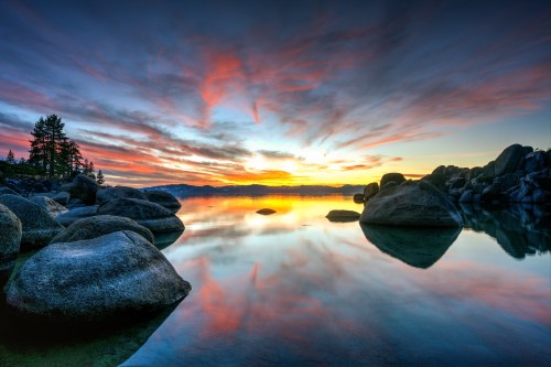 A serene lake at sunset, with colorful clouds reflecting on the water and large rocks lining the shore.