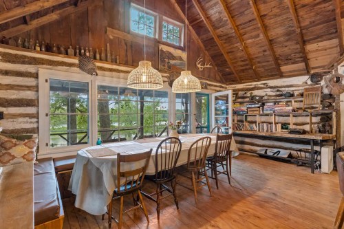 Cozy cabin interior with wooden beams, a large dining table, woven light fixtures, and a bookshelf by the window.