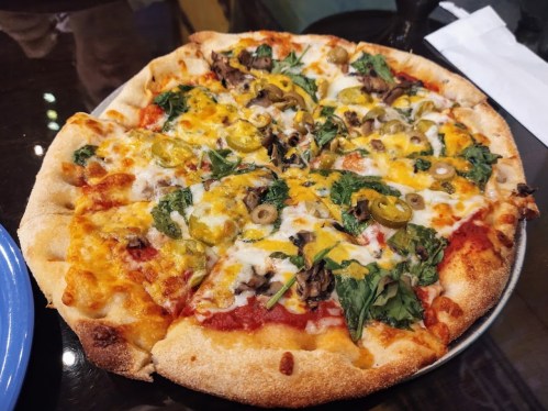 A freshly baked pizza topped with cheese, spinach, mushrooms, green olives, and jalapeños on a golden crust.