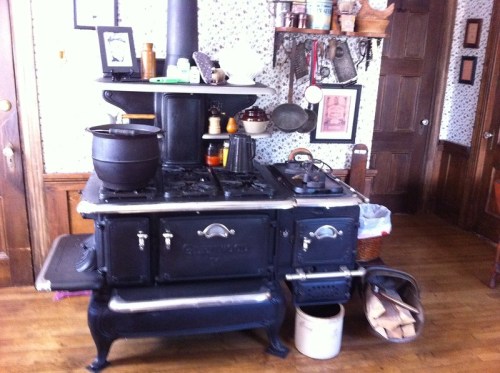 A vintage black stove with a pot on top, surrounded by kitchen utensils and decor in a rustic room.