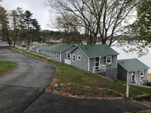 A winding road leads to a row of gray cabins with green roofs by a calm lake, surrounded by trees.