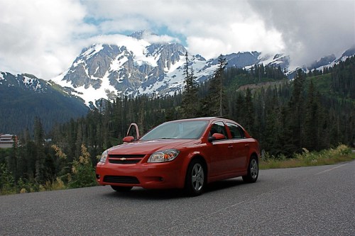 A red car parked on a scenic road with mountains and snow in the background under a cloudy sky.