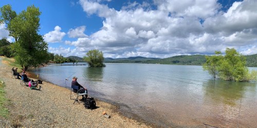 A panoramic view of a serene lake with people fishing along the shore under a partly cloudy sky.