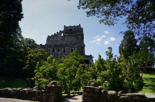 A stone ruin stands amidst lush greenery under a blue sky, framed by trees and colorful flowers.