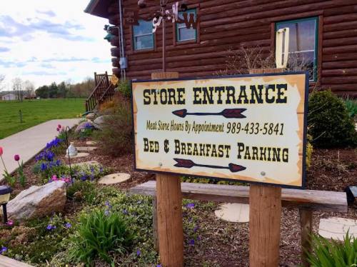 Sign at a log cabin entrance indicating store hours and bed & breakfast parking, surrounded by flowers and greenery.