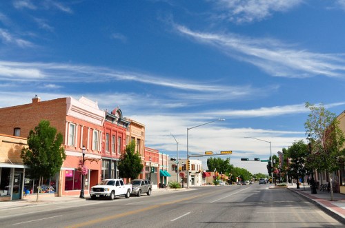 A quiet street lined with colorful buildings under a blue sky with scattered clouds. Trees and traffic lights are visible.