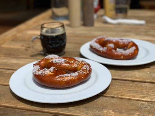 Two large pretzels with coarse salt on white plates, accompanied by a glass of dark beverage on a wooden table.