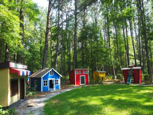 Colorful playhouses in a wooded area, surrounded by tall trees and green grass.