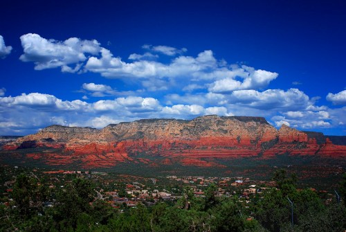 Vibrant red rock formations under a blue sky with fluffy clouds, overlooking a green valley and town below.